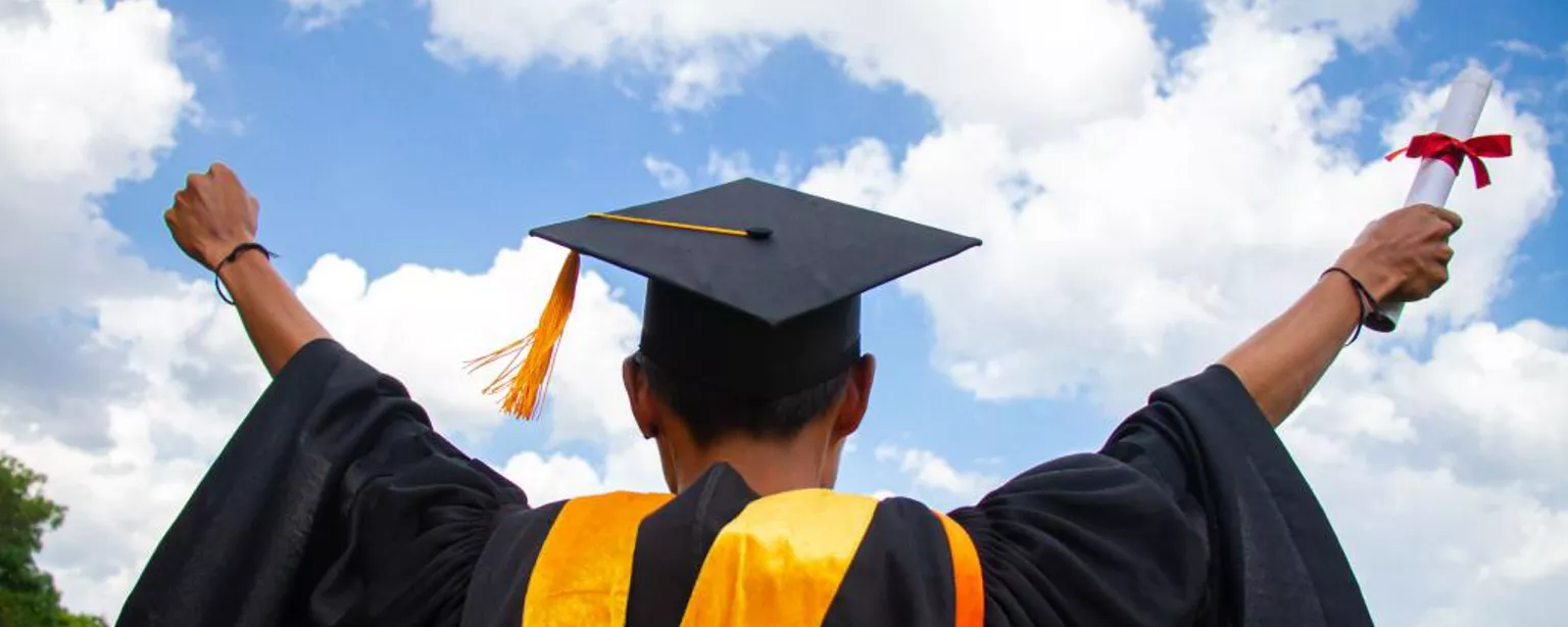 A college graduate with his back to the camera, arms outstretched to the sky and one hand holding a diploma.