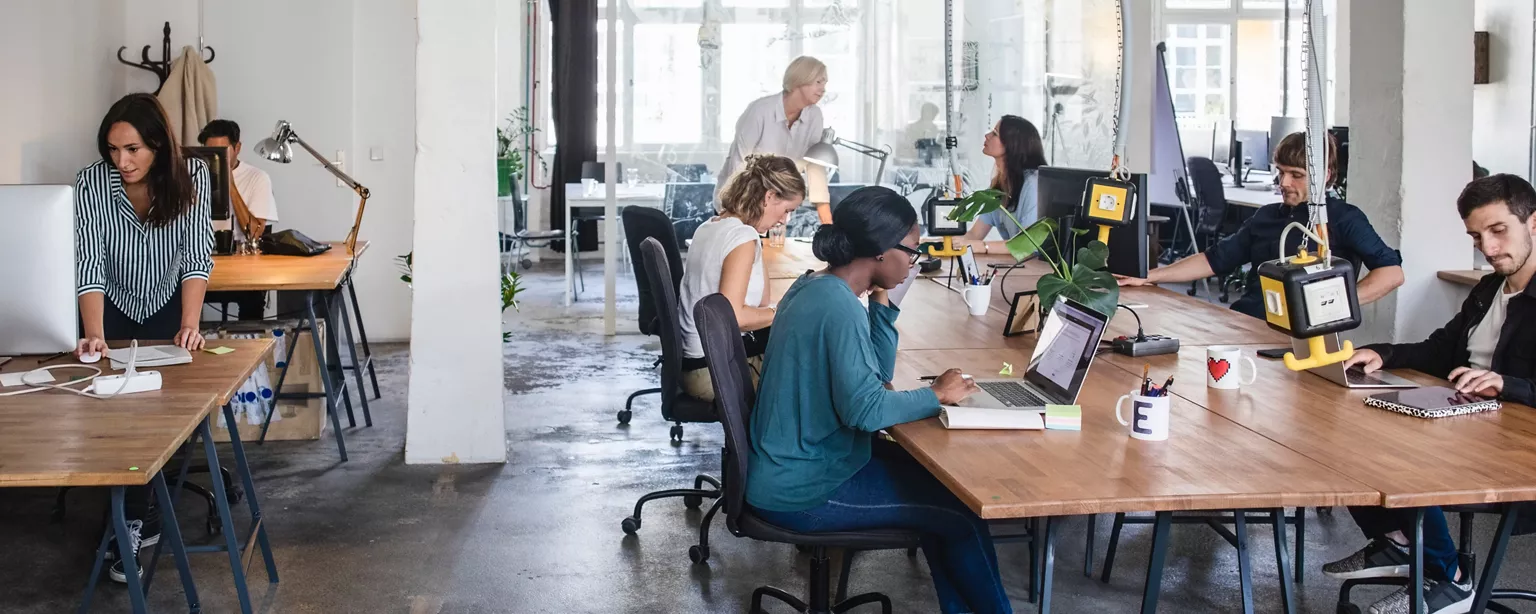 A group of colleagues work on their computers around a large wooden table in a modern industrial looking office.
