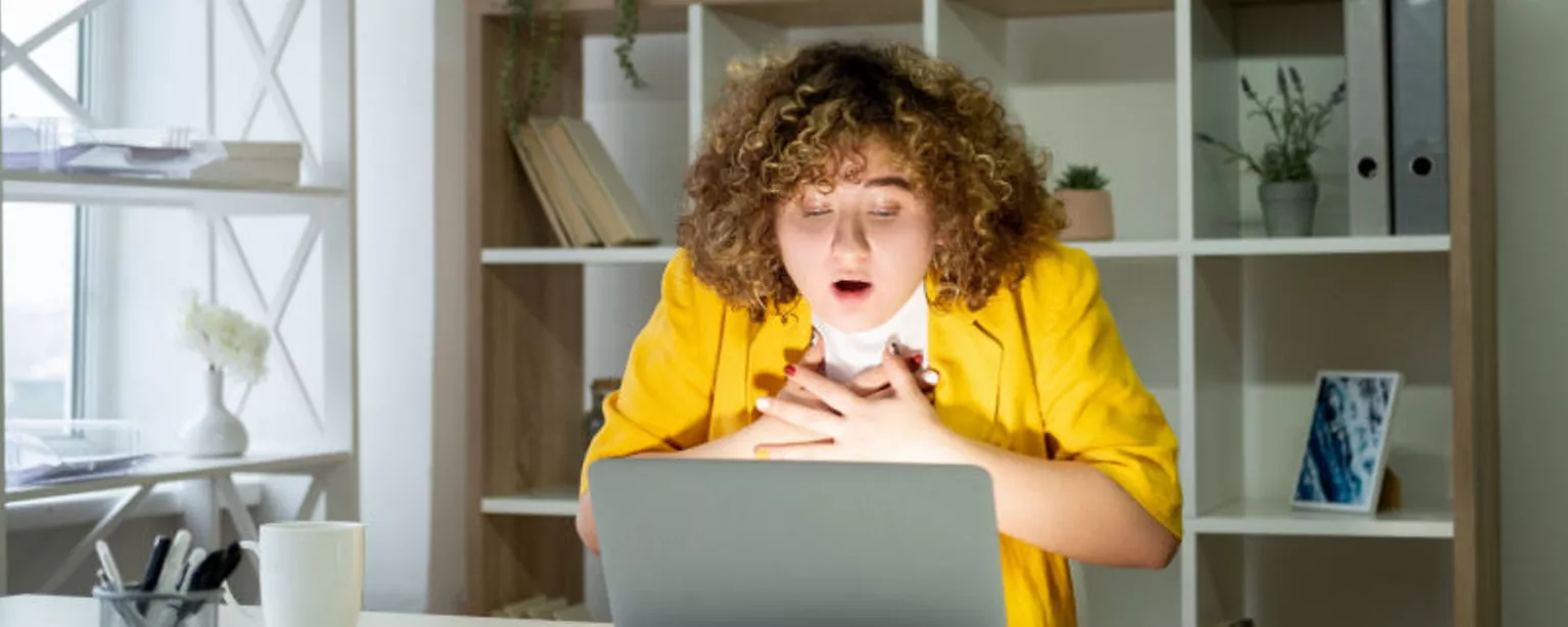 A woman who has fallen prey to an employment scam reacts by clutching her chest while sitting in front of her laptop.