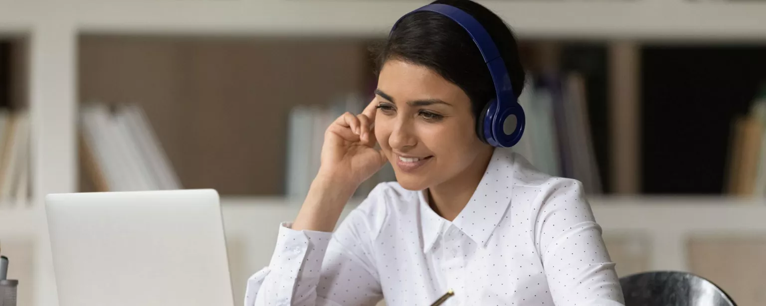 A smiling young woman wearing a white blouse with blue headphones, taking an online course and taking notes.