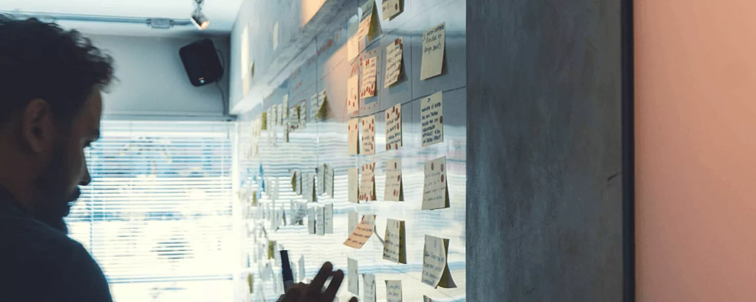Ein Mann steht mit Stift in der Hand vor einer Wand, an der sich viele Post-It-Notizen befinden