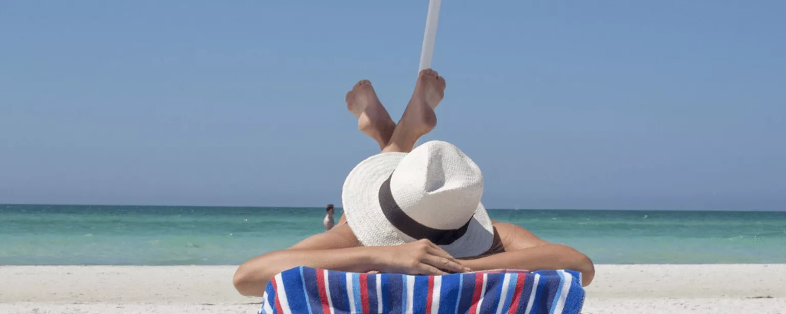 Person mit Hut über dem Kopf auf Sonnenliege am Strand.