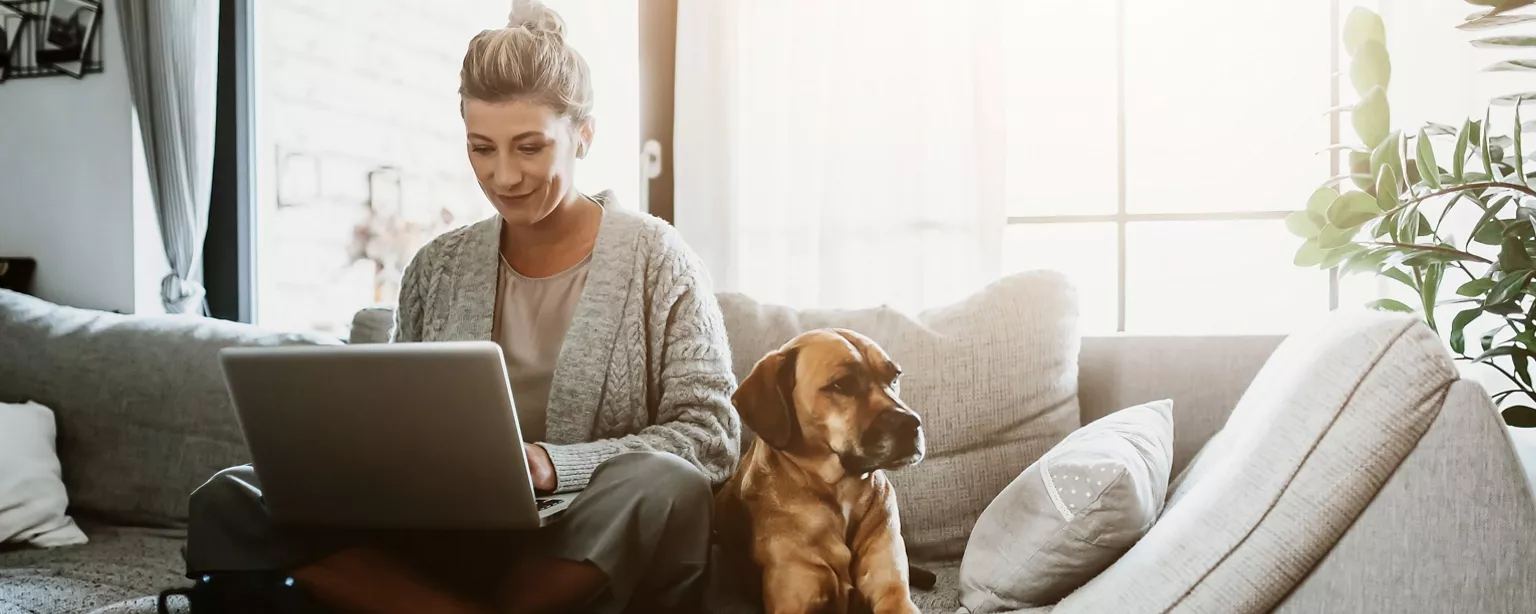 A woman works remotely from home as her dog sits next to her on a couch.