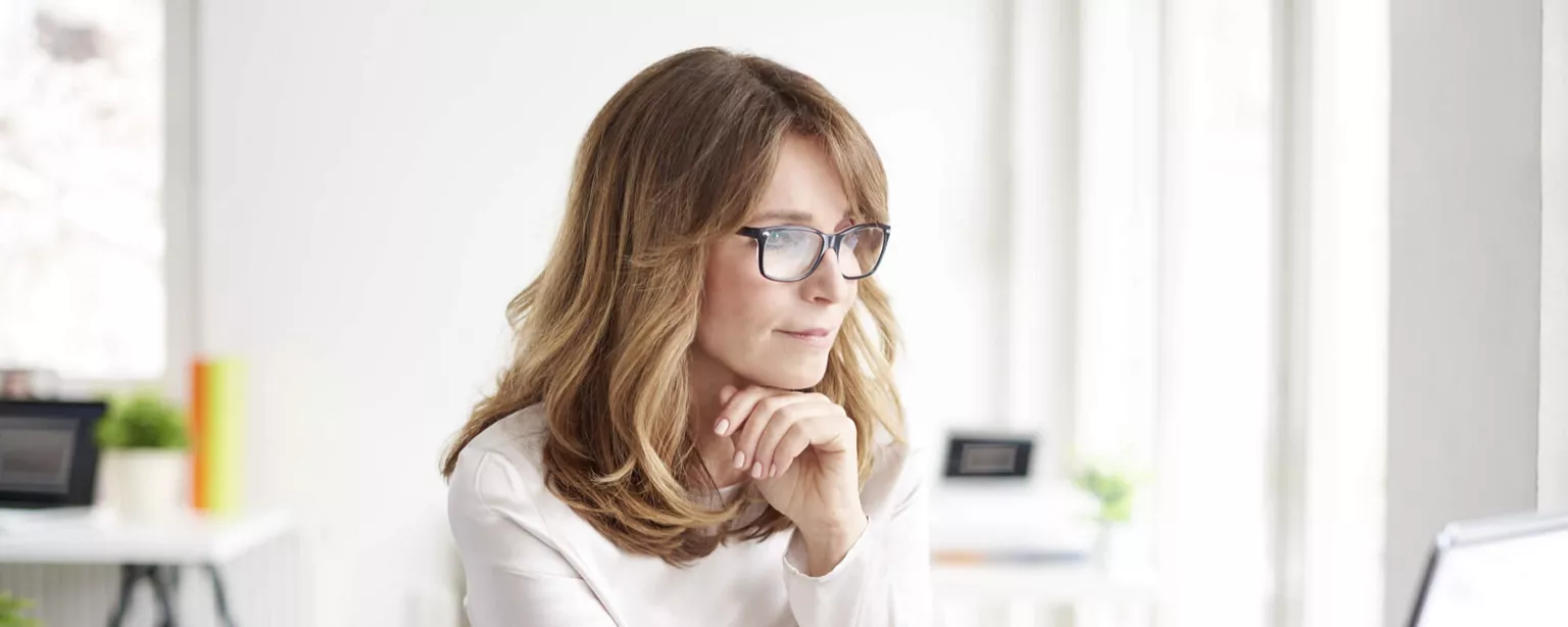 A woman wearing glasses, sitting at a desk in a home office and working on a laptop.