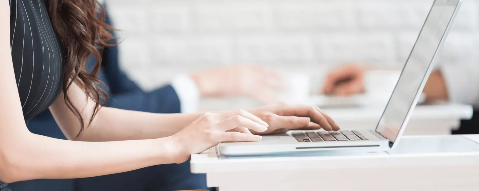 A young woman typing at a desk on a laptop with other workers in the background.