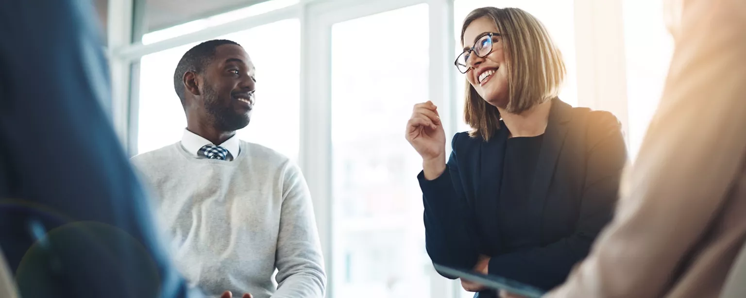 A man and a woman sit in front of windows talking in a group setting.