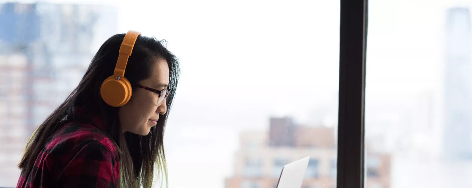 A woman in a red checked shirt with yellow headphones works on a laptop