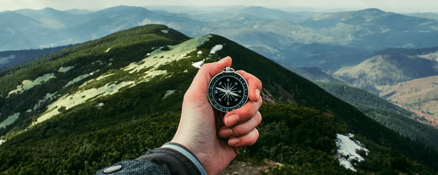 Eine Hand hält vor einer Gebirgskulisse einen Kompass hoch