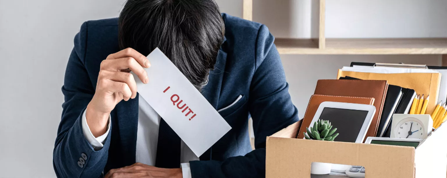 Ein Businessman sitzt erschöpft am Tisch, lässt den Kopf hängen und hält einen Zettel mit der Aufschrift "I Quit".