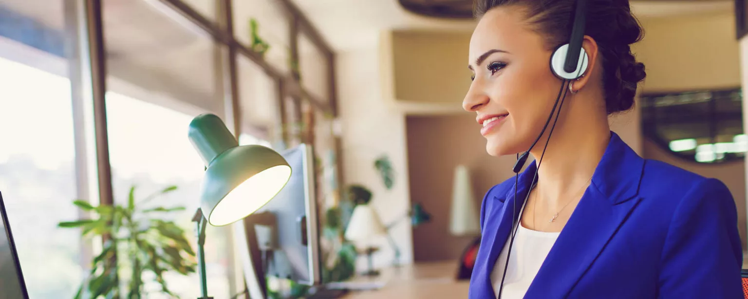 A smiling, professional woman appears happy with her career as she talks on a headset.and works at a computer.