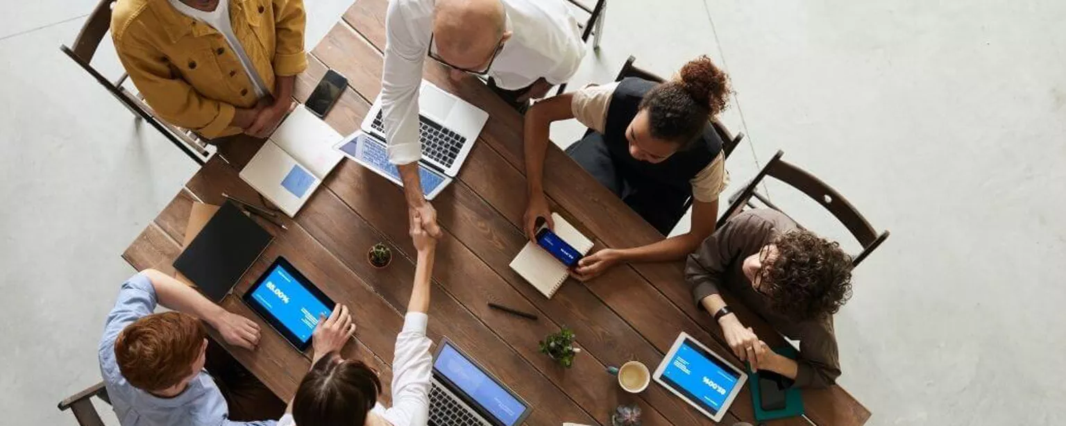  In einer Runde mit mehreren Beschäftigten besiegelt der Leiter per Handschlag quer über den Tisch die Aufgabenverteilung an ein Teammitglied  