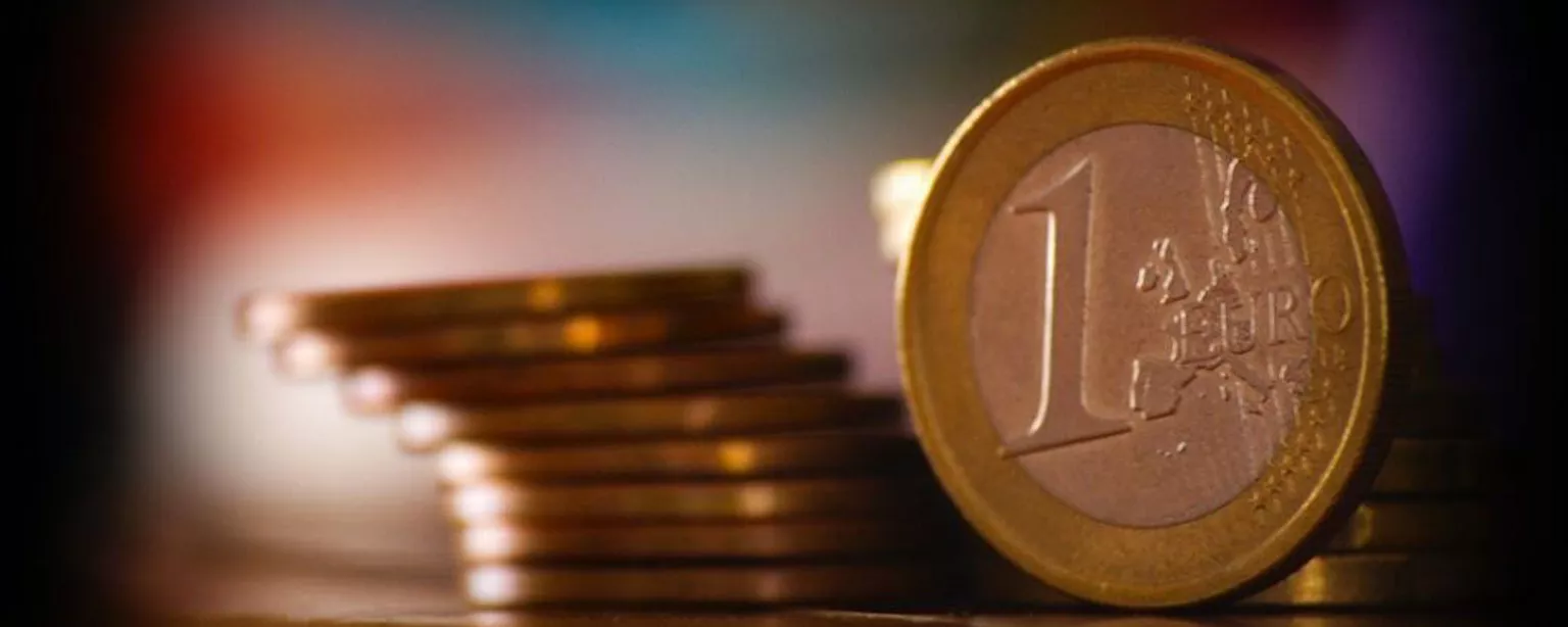 Ein Stapel Münzen liegt auf einem Tisch, im Vordergrund eine aufgestellte 1-Euro-Münze  
