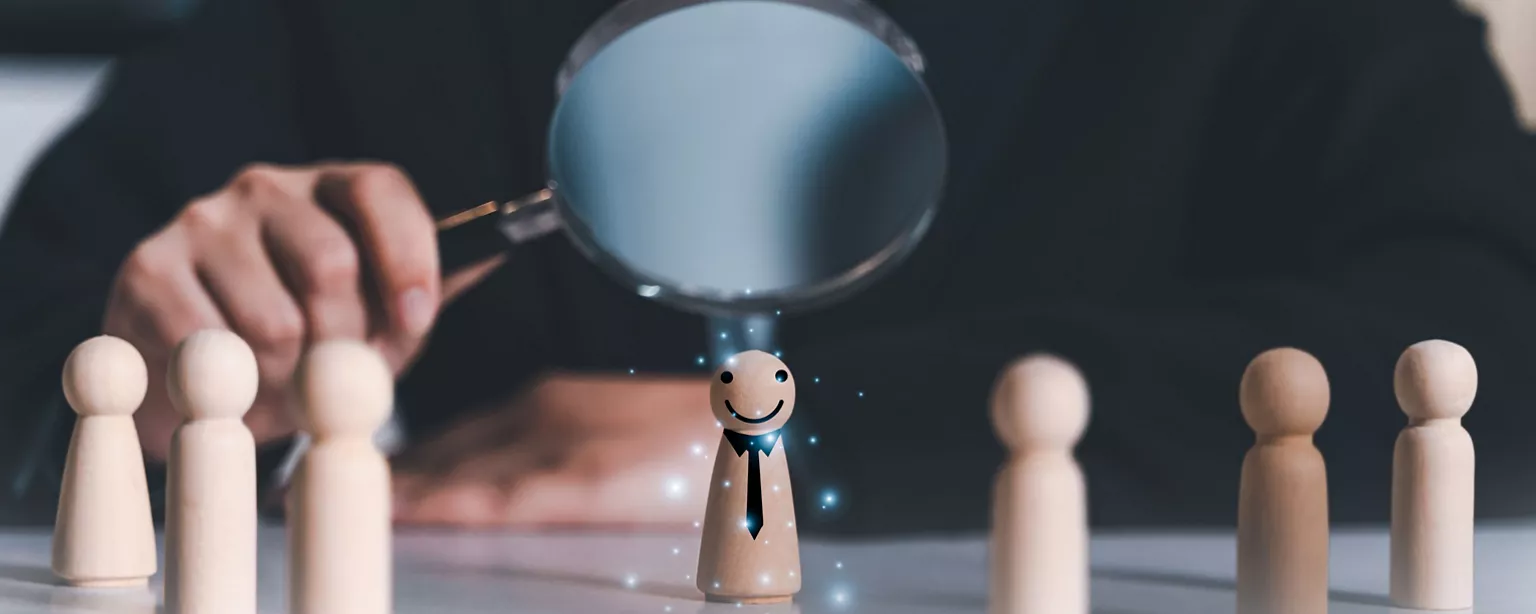 A hand holds a magnifying glass to highlight a wooden figure representing a nontraditional candidate.