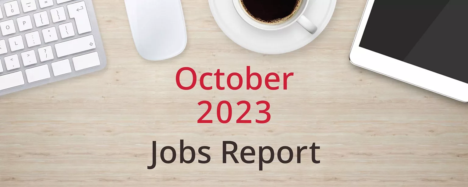 On a wooden desktop, positioned below a keyboard, mouse, cup of coffee and a tablet computer, are the words, “October 2023 Jobs Report.”