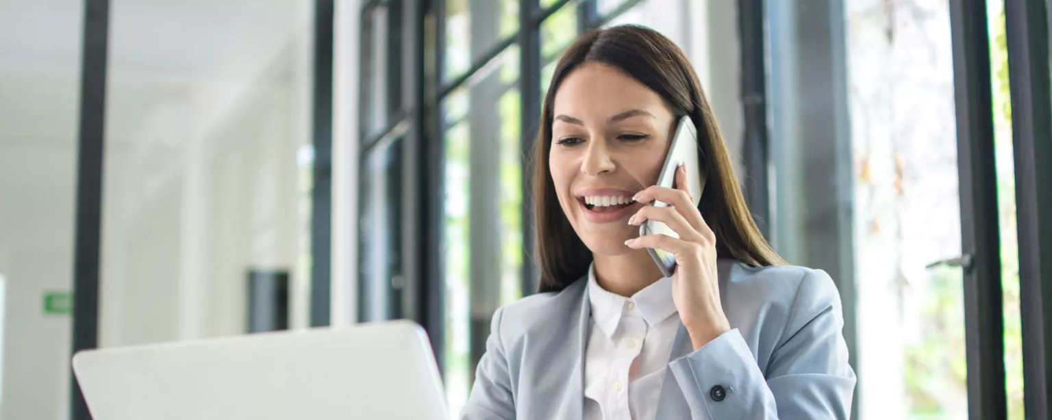 Young woman in blazer sitting at desk in an office in front of a laptop, talking on mobile phone and smiling.