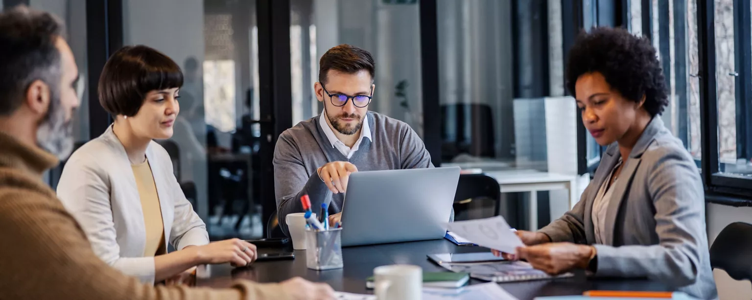 A project team leader discusses work status with a team of project professionals in a conference room.
