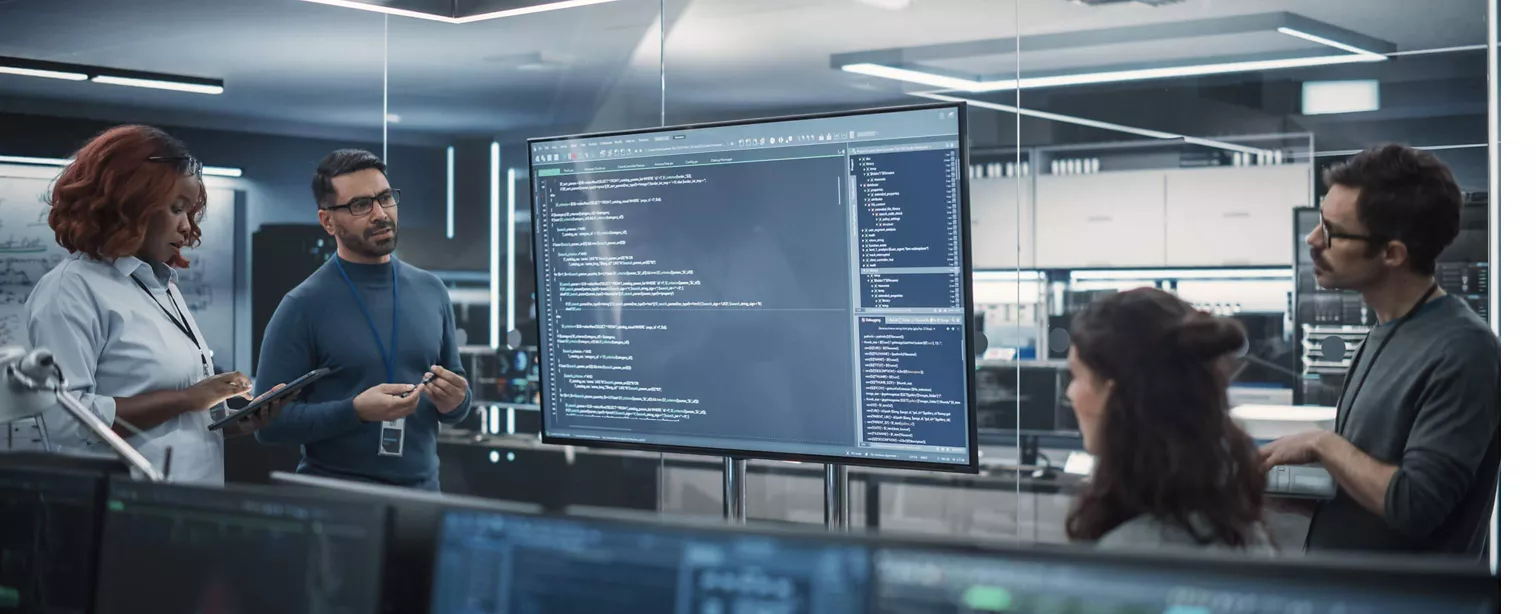 A technology leader discusses an AI initiative with his team; they are all standing around a large screen displaying computer code.