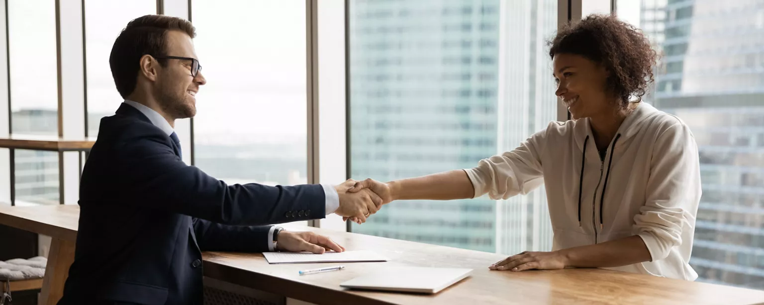 A staffing agency recruiter and a job seeker shake hands across a table in a high-rise office.