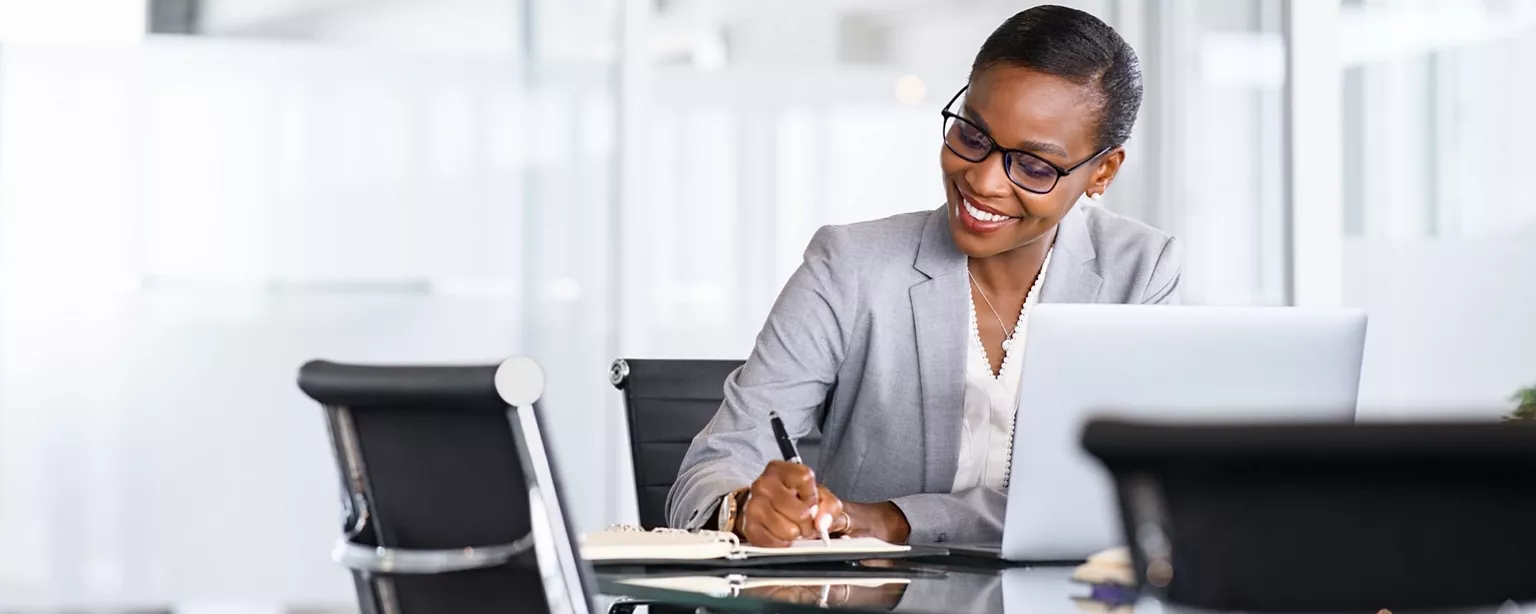 A smiling and sharply dressed woman in finance outlines her career goals in a notebook; she is sitting at a conference room table with a laptop open in front of her.