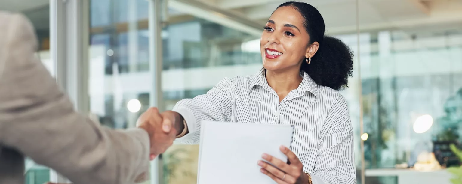 A hiring manager greets a prospective employee with a smile while holding the applicant's resume.