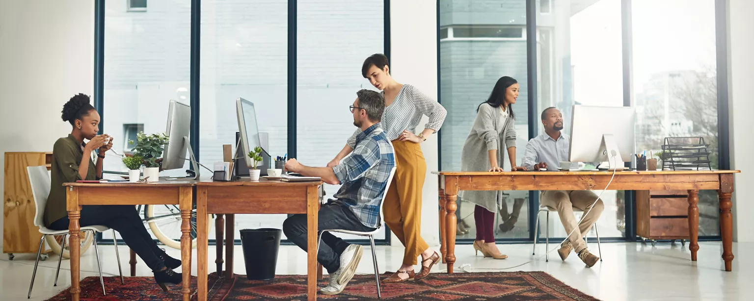 Employees work in a modern office. Some workers are collaborating while looking at computer screens, while one works independently.