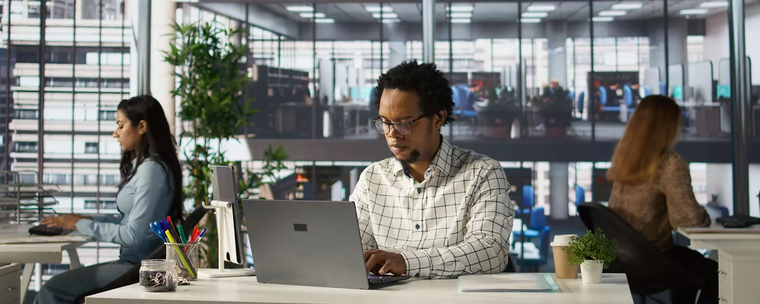 Three employees work at laptops in a modern office; they are ssitting at workstations facing different directions.
