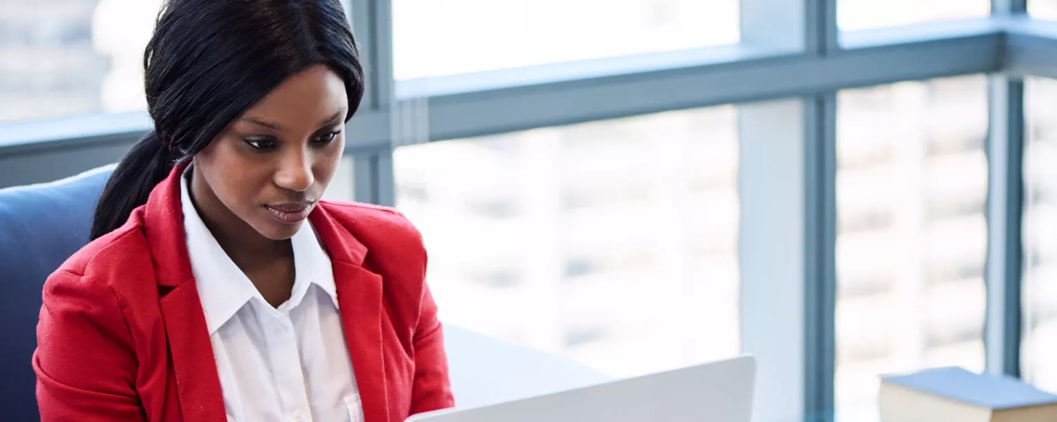 Woman in a red blazer and white shirt working on a laptop in a high-rise office.