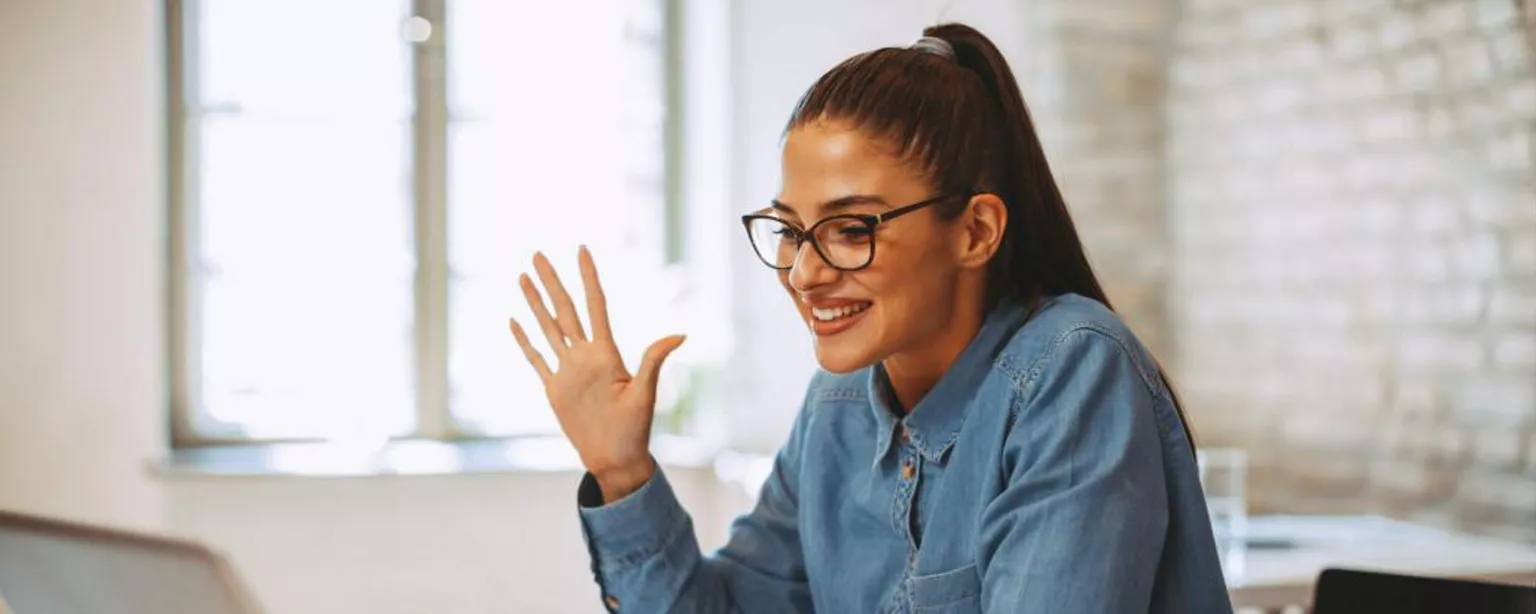 Young woman smiling, sitting at a table in front of a laptop computer, waving at her screen.