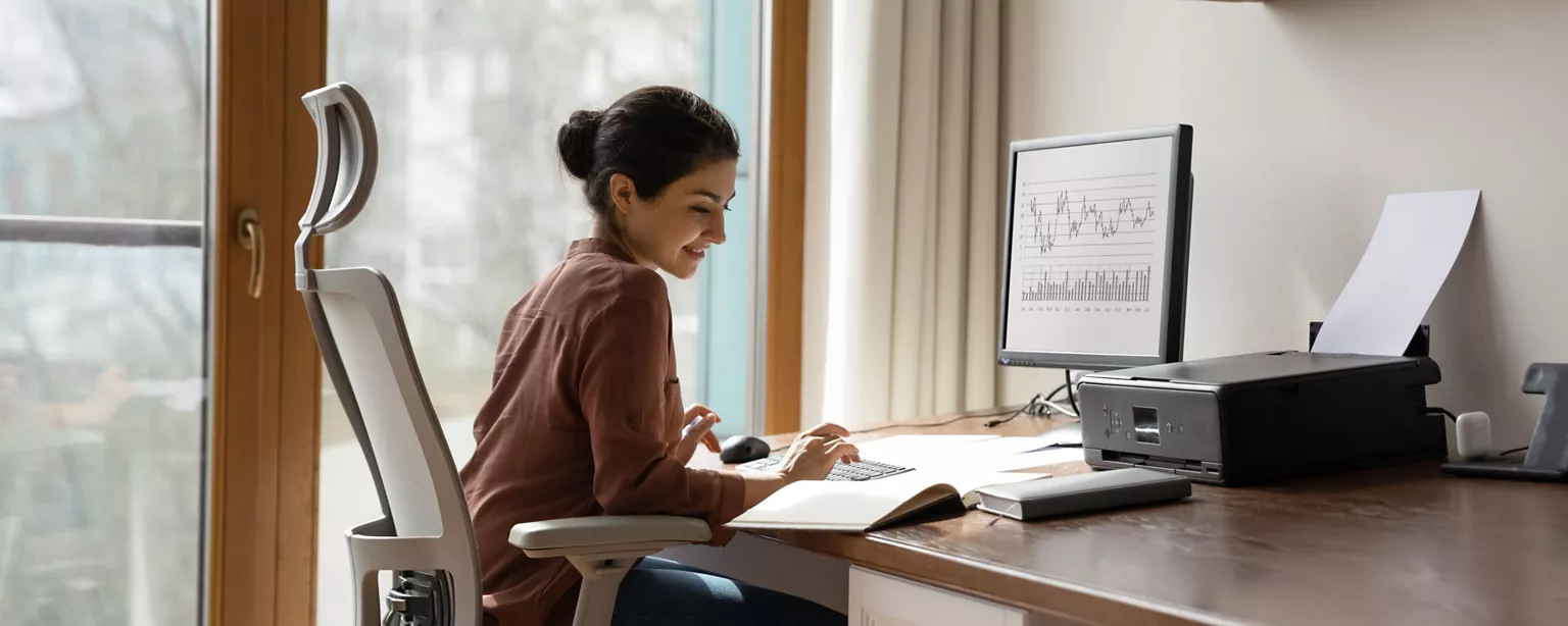 A woman in a brown shirt sits in her home office in front of a computer and printer; she is smiling as she works on a project.