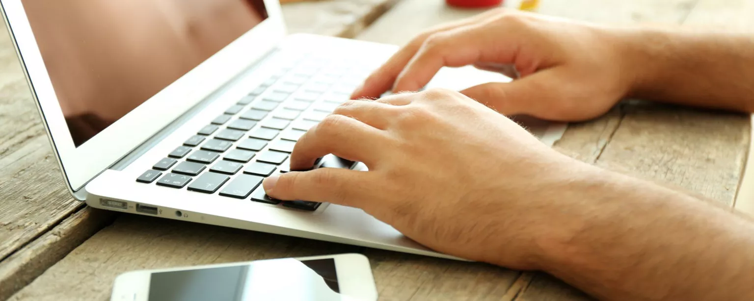 A man's hands typing on a laptop on a wooden desk with a mobile phone and red coffee cup nearby.