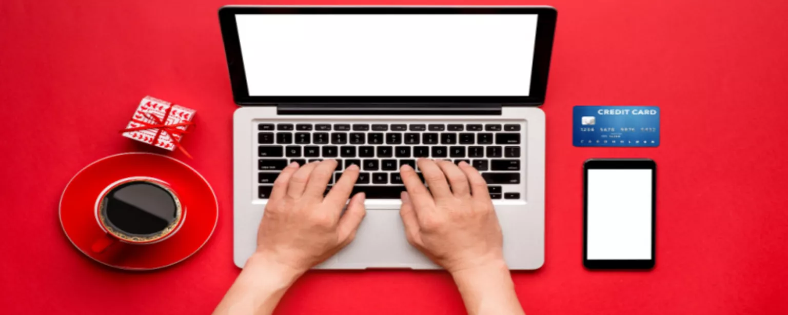 Overhead view of man's hands typing on laptop against red background surrounded by mobile phone, credit card, small wrapped present and cup of coffee