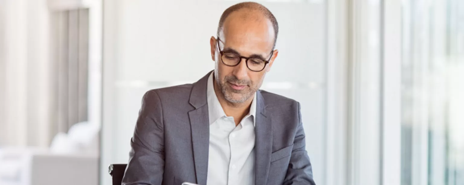 A businessman in glasses and a gray blazer sitting at a desk in a corporate office and looking at his smartphone.