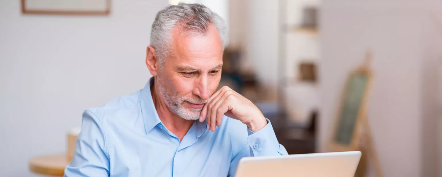 An older man with gray hair and a beard is thoughtfully looking at a laptop screen while looking for retirement jobs.