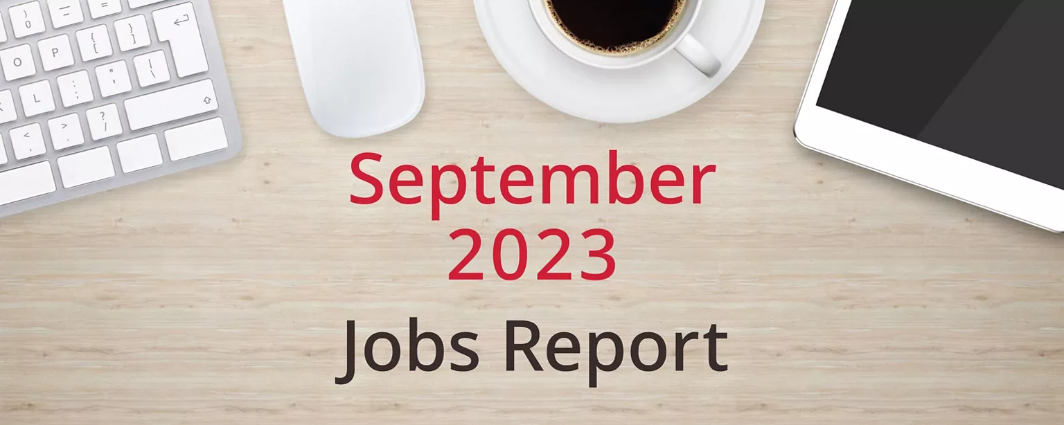 On a wooden desktop, positioned below a keyboard, mouse, cup of coffee and a tablet computer, are the words, “September 2023 Jobs Report.”