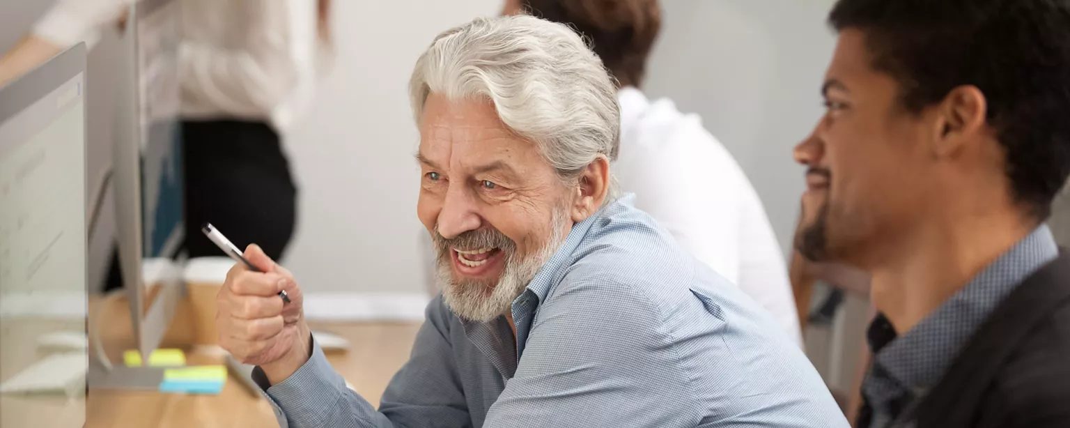 A mature business man sits at a desk in an office next to a colleague and both are looking at the same computer as if working together on a project.