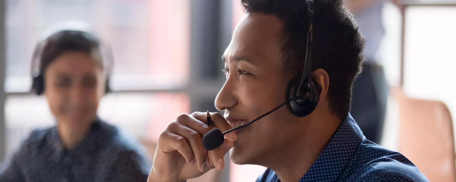 A smiling young businessman talks on headset as a smiling coworker looks in his direction.