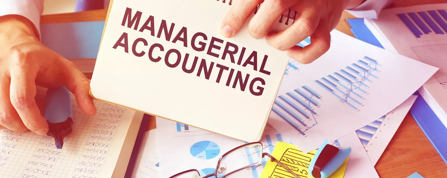 A sign that says Managerial Accounting is on a desk with a hand holding it and financial paperwork in the background.