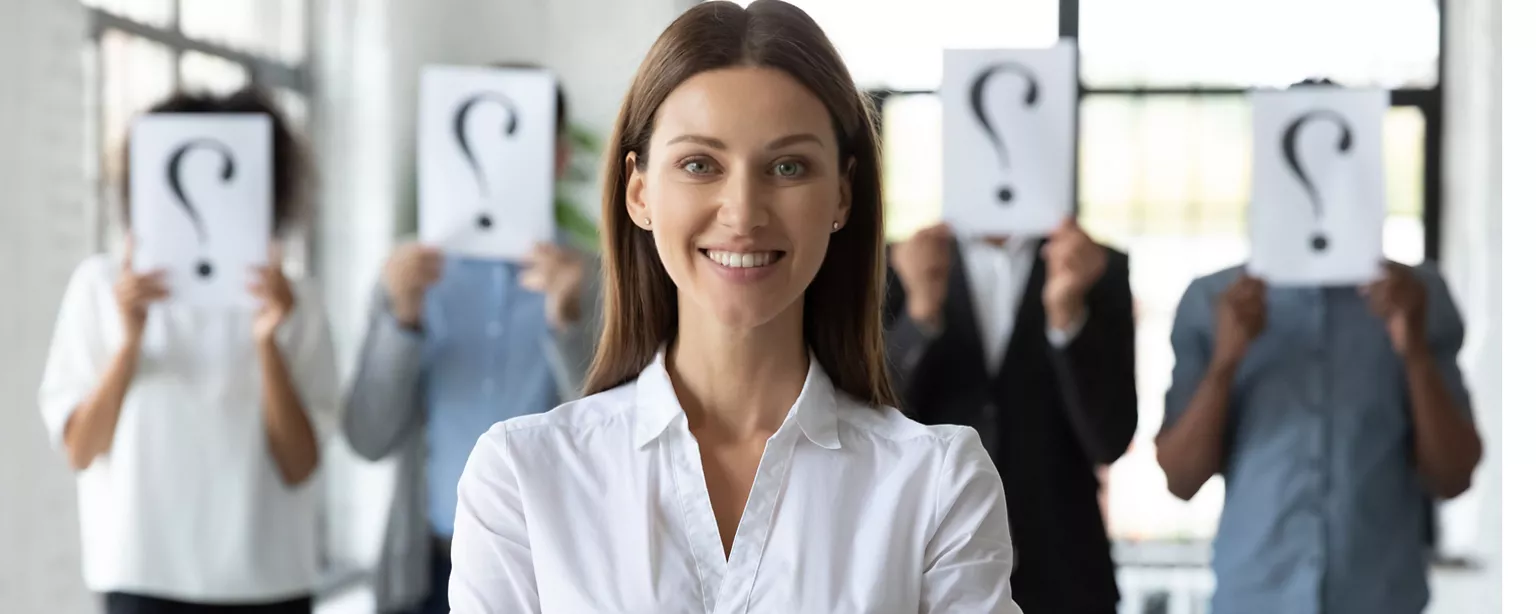 Confident woman in white blouse standing in front of four other job applicants holding a piece of white paper with a question mark over their faces.