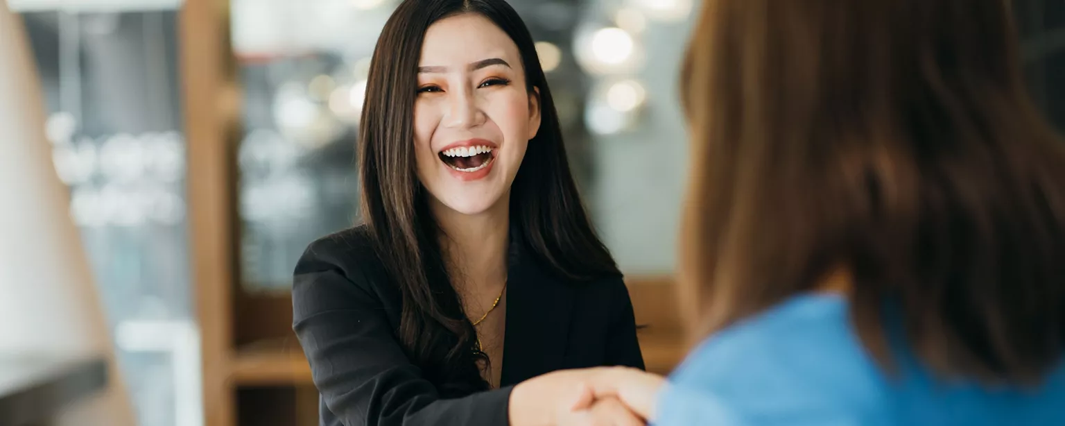 Job candidate smiling and wearing black blazer shaking the hand of an interviewer.