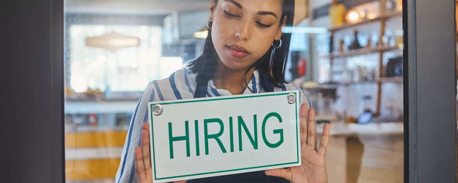 A small business owner posts a "HIRING" sign in her storefront window.