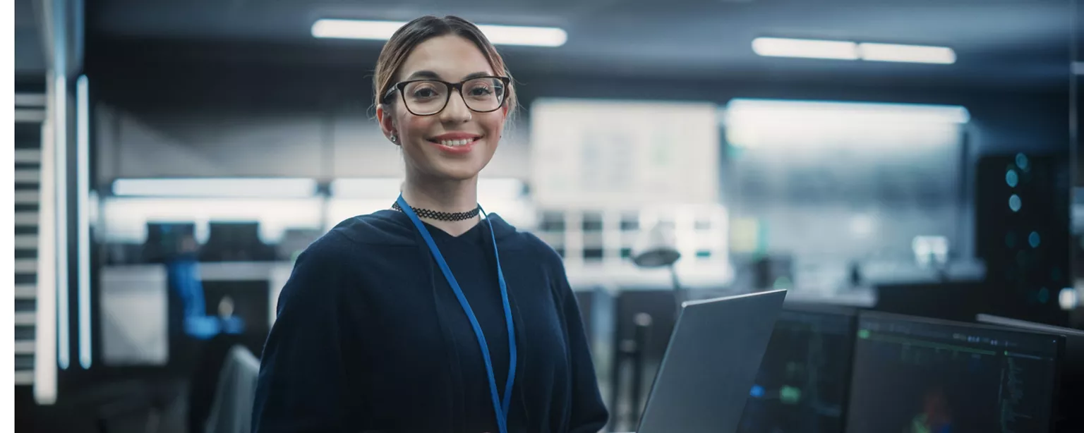 An IT project manager smiles while holding a laptop at work.