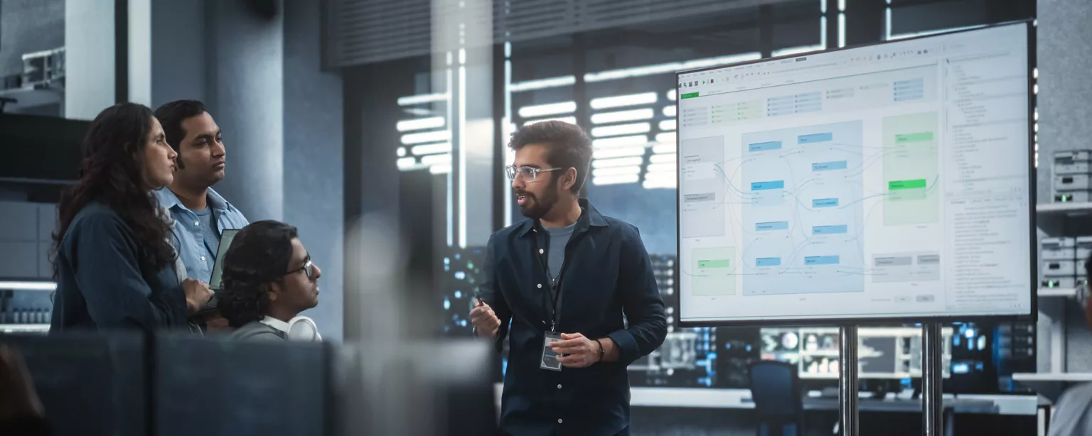A technology leader discusses an AI initiative with his team; they are standing and sitting around a large screen displaying charts and words..