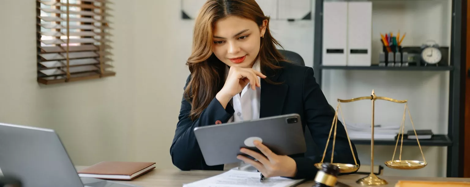 Female legal professional seated at desk viewing digital content on a tablet, with model of scales in the foreground