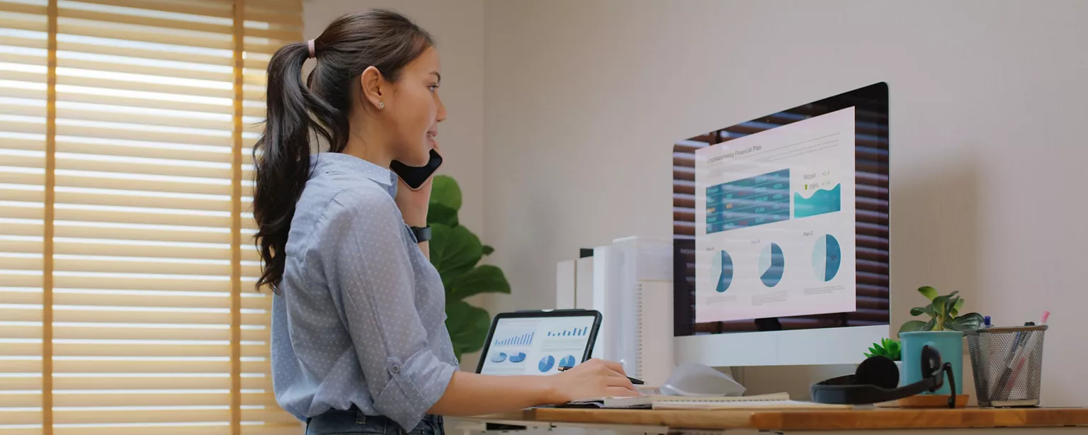 A woman in a blue dress shirt stands in her home office in front of a computer; she is staring at pie charts and talking on the phone.