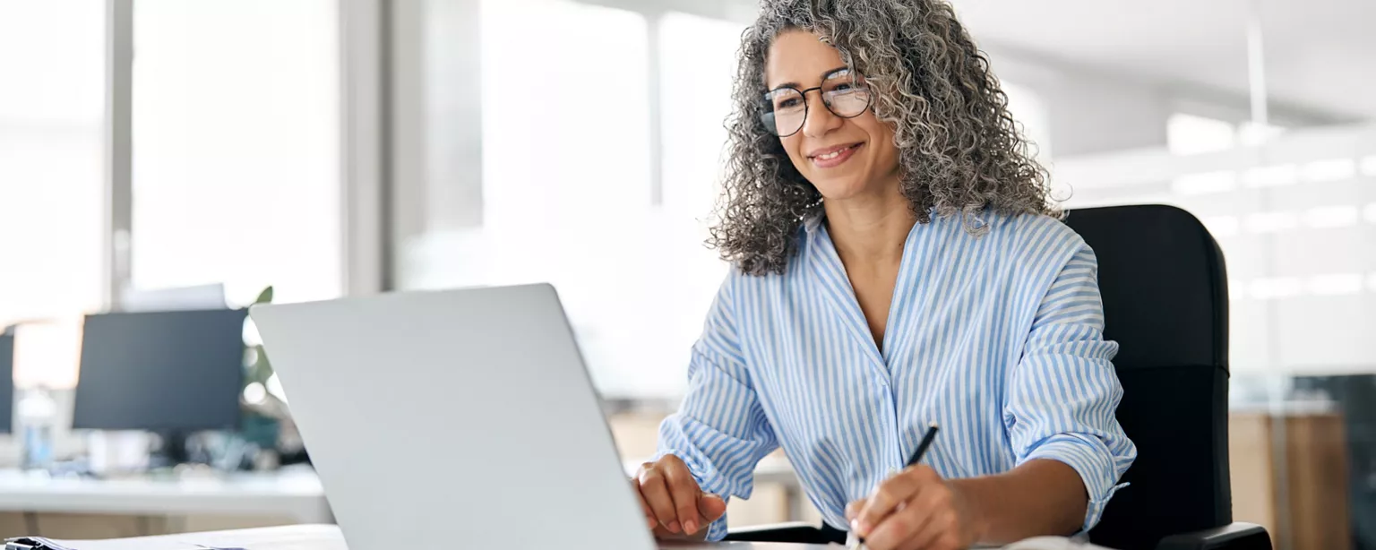 A smiling professional woman with curly gray hair works on a laptop and takes notes at her desk in an office.