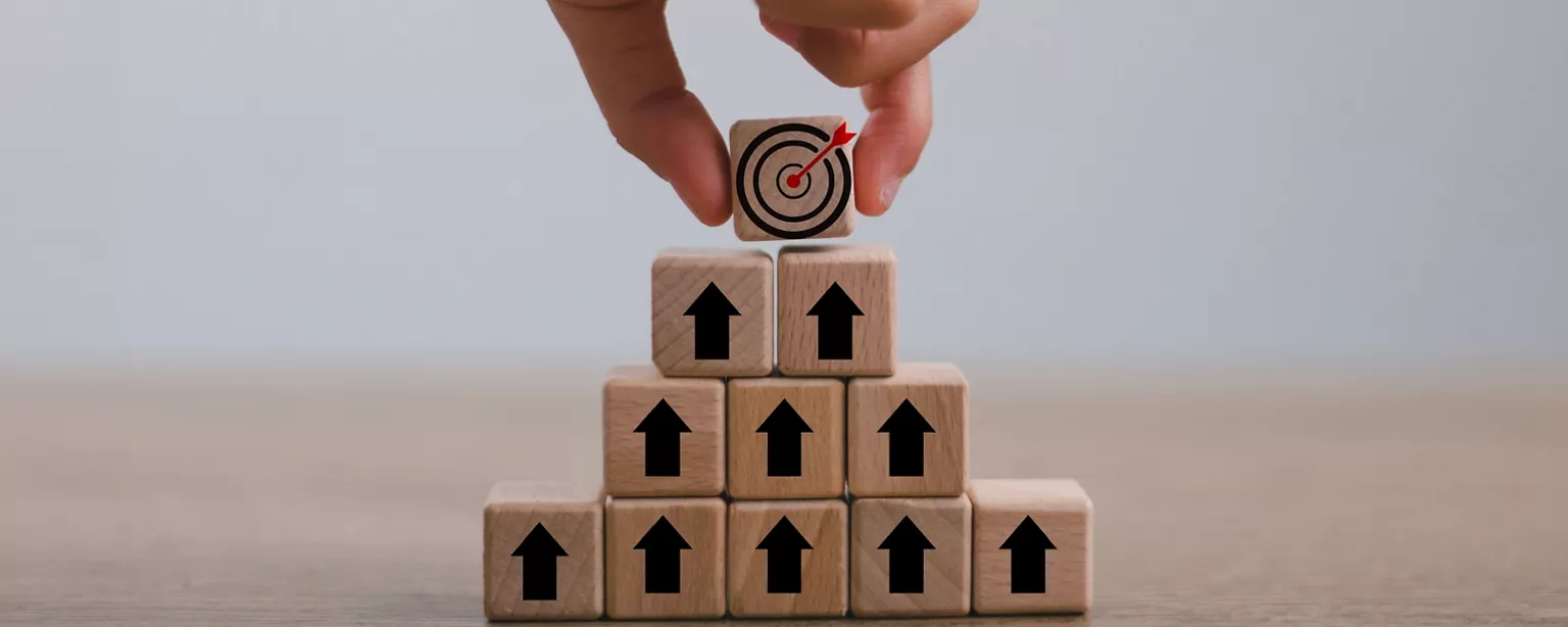 A pyramid of square blocks with an arrow on each pointing up and a hand putting the final block on top with a bullseye.