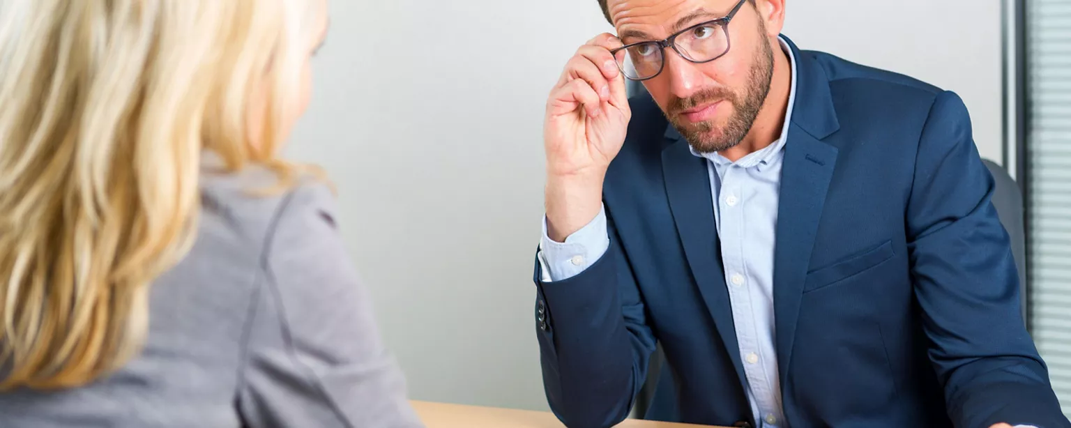 A hiring manager holding a candidate's resume casts a skeptical eye as he speaks during a job interview.