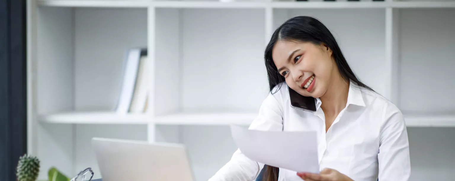 A smiling woman working a small business is holding a sheet of paper while looking at a laptop.