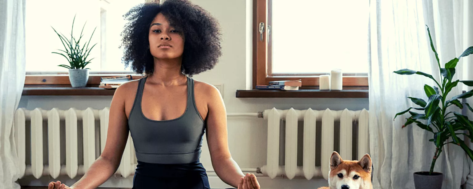 Eine junge Frau sitzt mit geschlossenen Augen im Lotussitz auf ihrer Yoga-Matte, neben ihr sitzt ihr Hund
