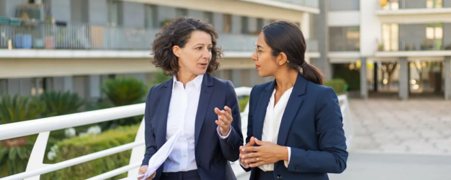 Two businesswomen, a mentor and a mentee, walking and talking outside in an office complex courtyard.